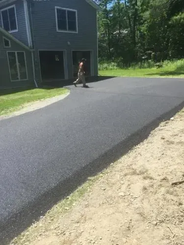 Newly paved asphalt driveway leading to a two-story gray house. A person walks on the driveway.