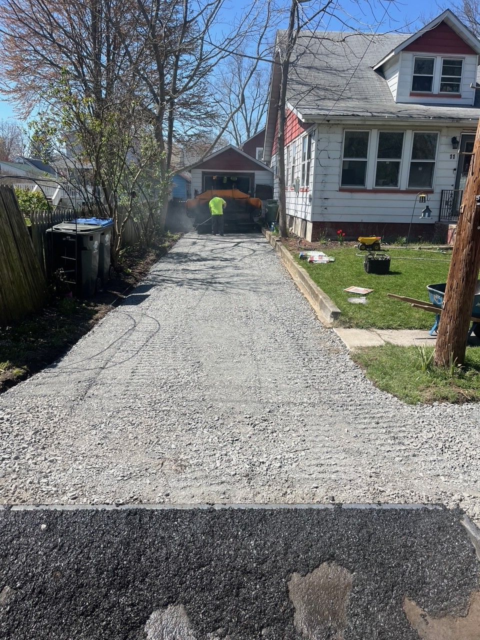 Driveway under construction: gravel base with a person and equipment in the distance; houses and trees are in the background.