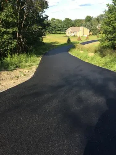 Black asphalt driveway curves toward a beige house in a green, rural landscape.