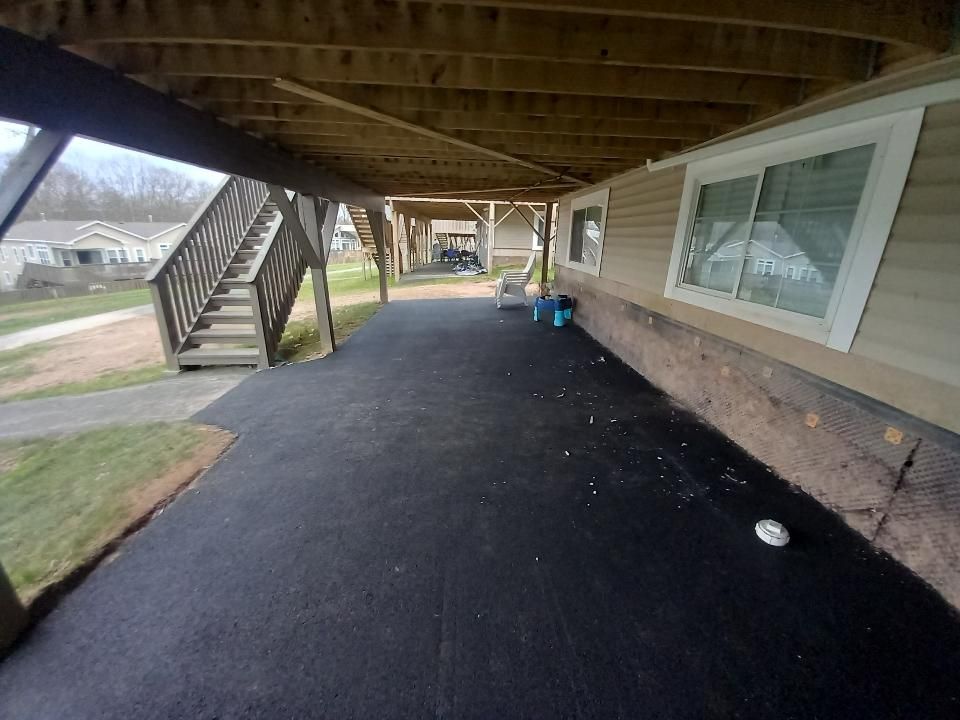 Underneath a raised structure, a dark asphalt patio extends toward wooden stairs, with a light-colored building to the right.