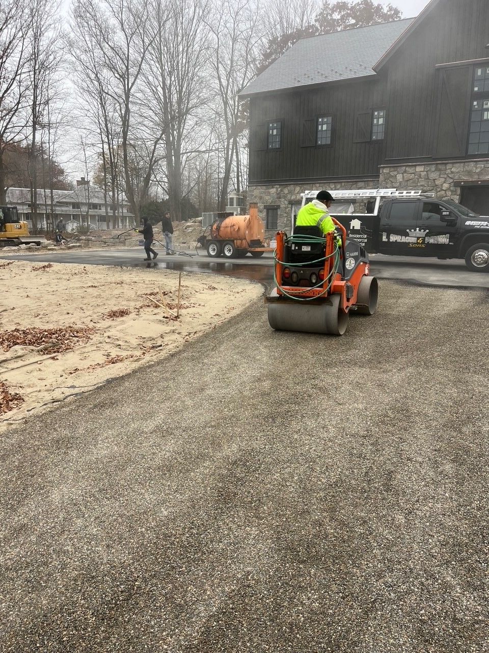 Man operating a roller compactor compacting gravel on a construction site near a building.