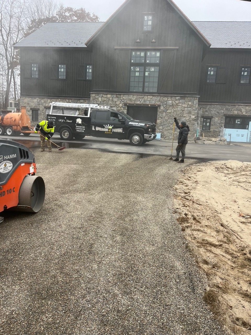 Construction workers paving a gravel driveway in front of a large dark-colored building.