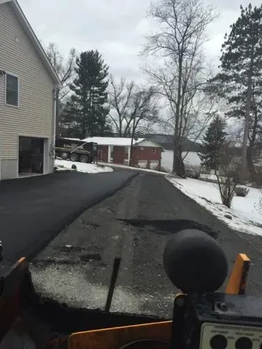 Asphalt driveway being paved, snow on the side, house on left, water in background.