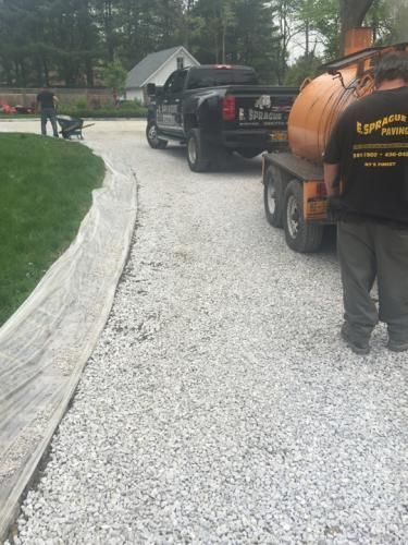 Truck and trailer on a gravel driveway, workers nearby. Driveway is bordered by grass and landscaping.