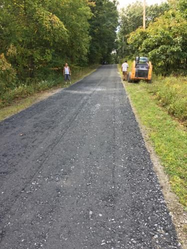 Newly paved road with two people standing on the side, and a skid steer. Trees line both sides.