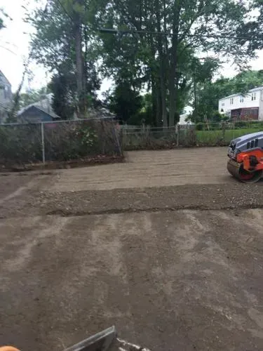 A compacted dirt lot with a roller, trees, and a fence.