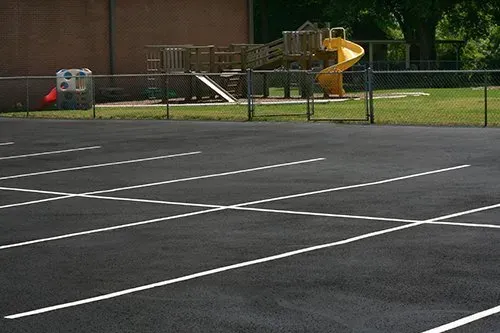 Parking lot with white lines in front of a fenced playground with slide.