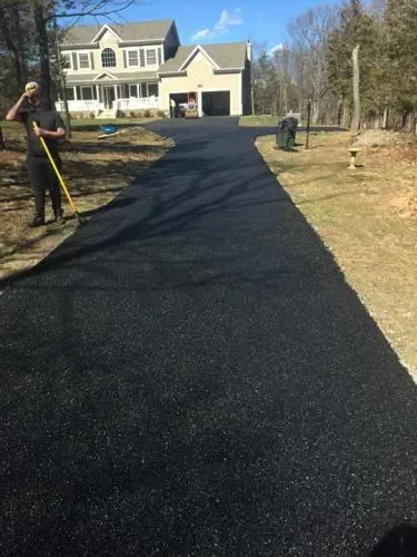Newly paved black asphalt driveway leading to a two-story house with a garage. Man with rake stands near driveway.