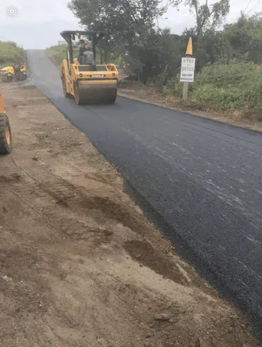 Road roller compacting fresh asphalt on a country road. Yellow machinery and dirt shoulder visible.