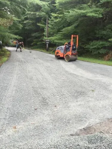 Gravel road being compacted by a small roller; a person shovels nearby, trees line the road.