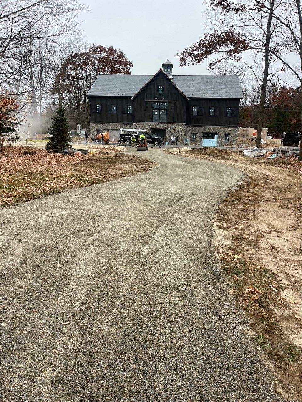 Gravel driveway leading to a dark, two-story barn under construction, surrounded by trees.