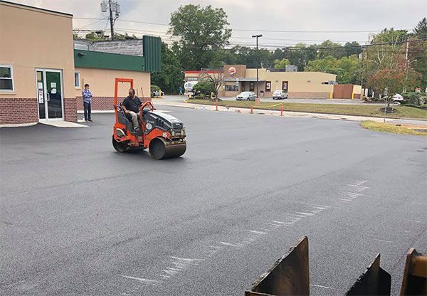 Man operating an orange asphalt roller on a freshly paved parking lot. Buildings in the background.