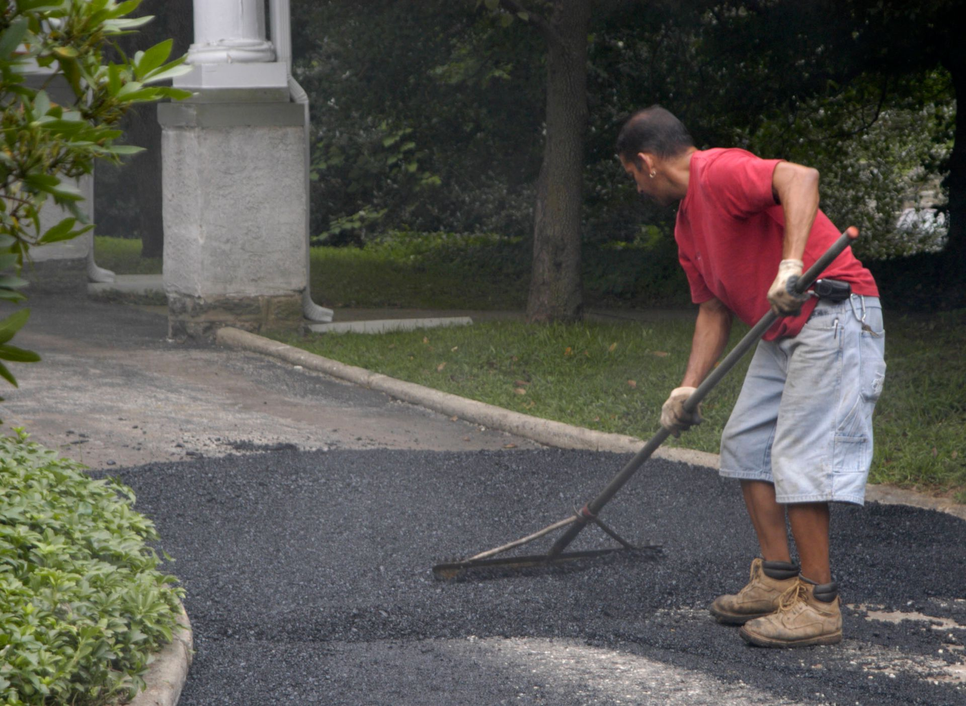 Man raking asphalt on a driveway next to a white pillar and greenery.