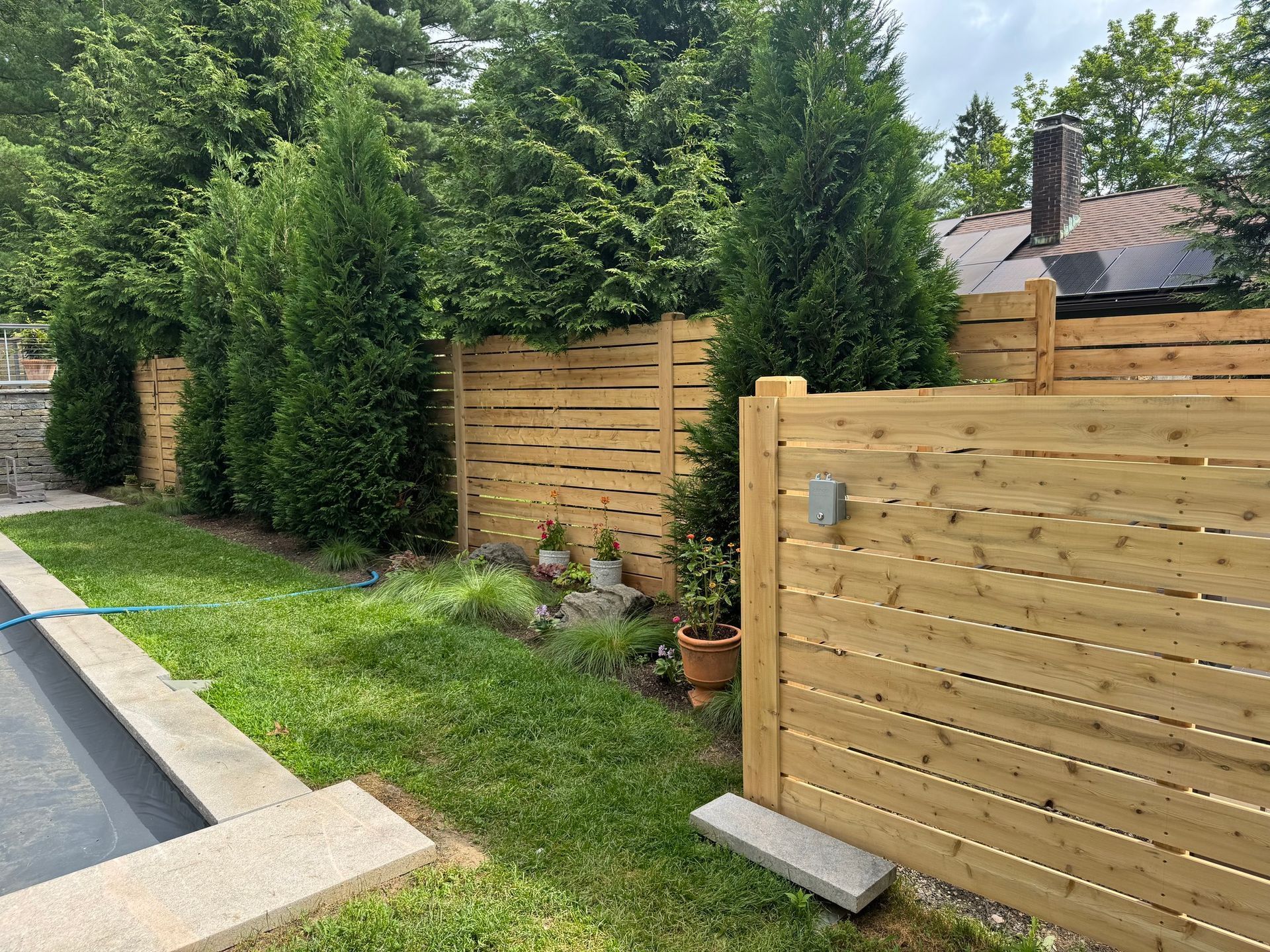 A wooden horizontal slat fence borders a yard with green grass and evergreen trees.