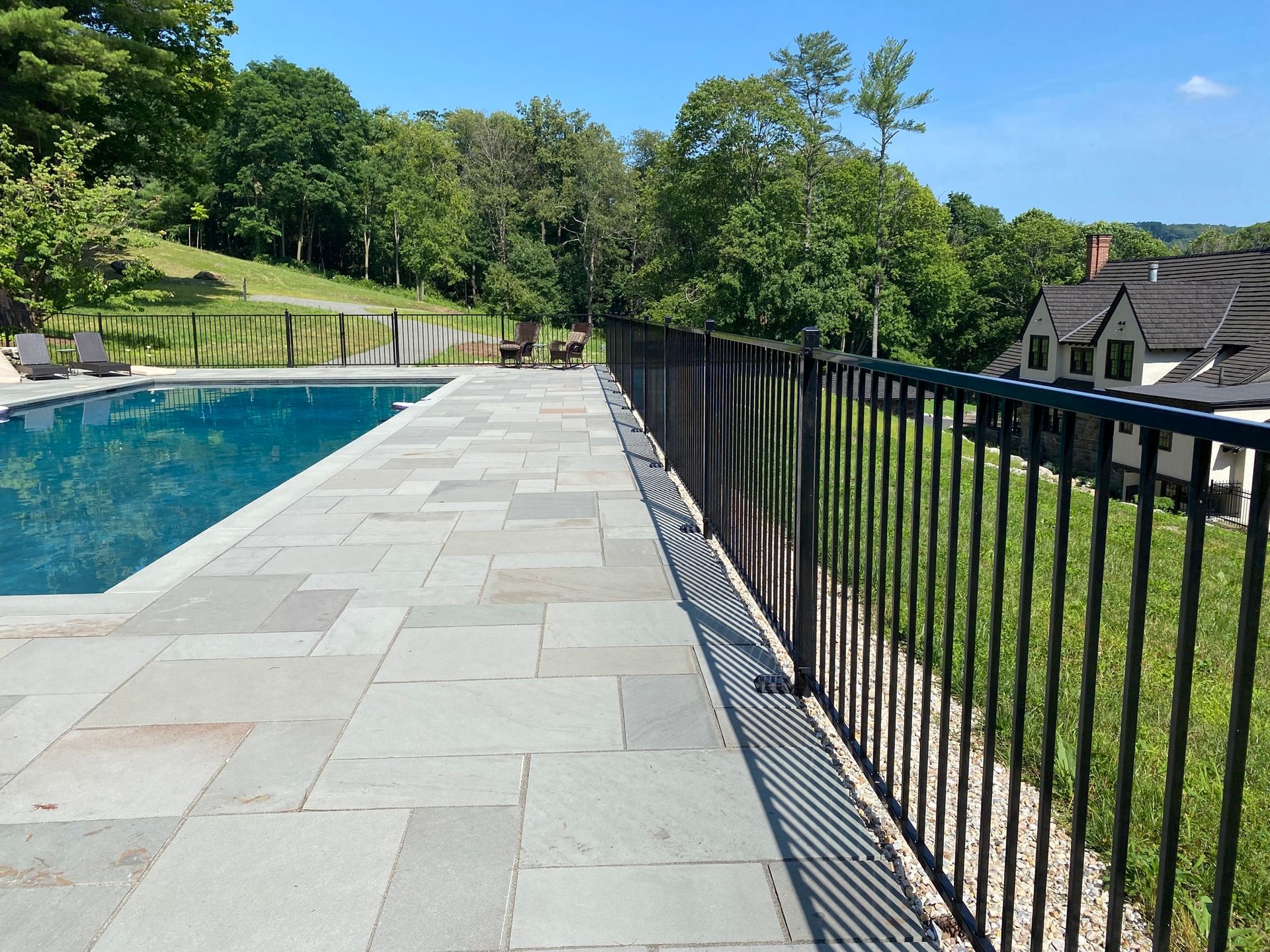 Poolside view with a black fence, stone patio, and a house in the background on a sunny day.
