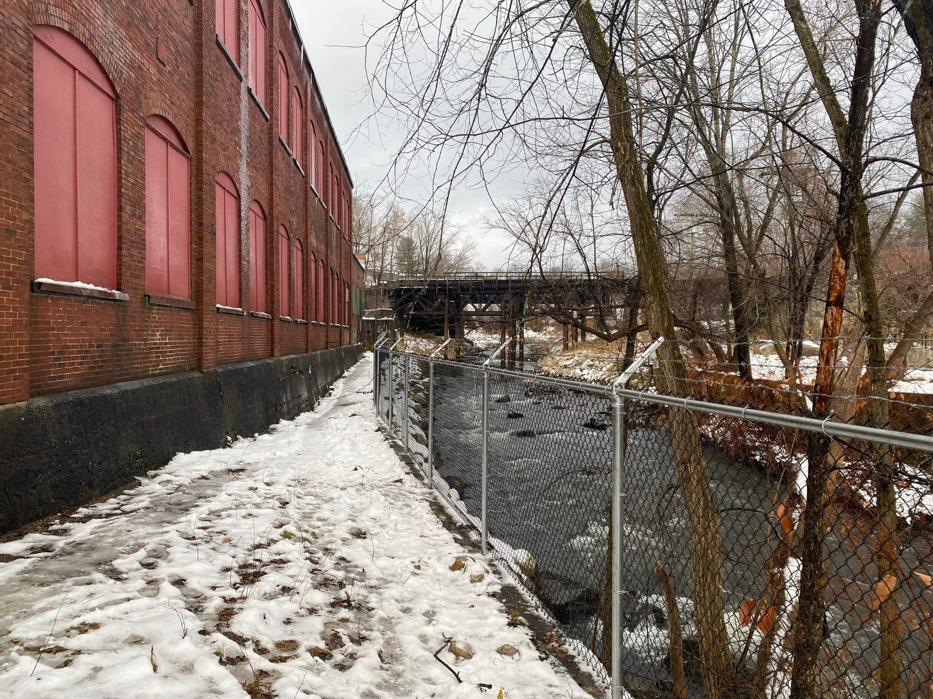 Brick building next to a snowy path and a river, separated by a chain-link fence.