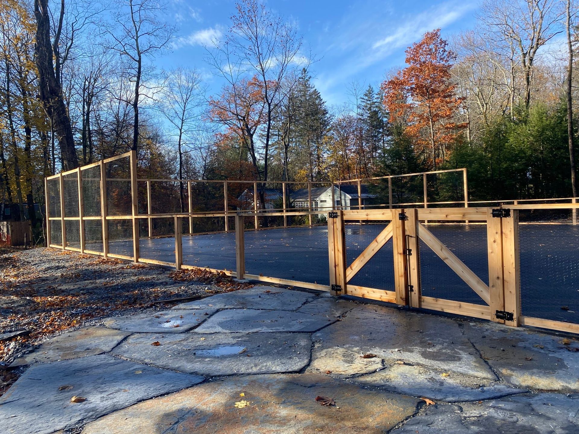 Wooden fence and gate enclosing a paved area, surrounded by trees with autumn foliage.