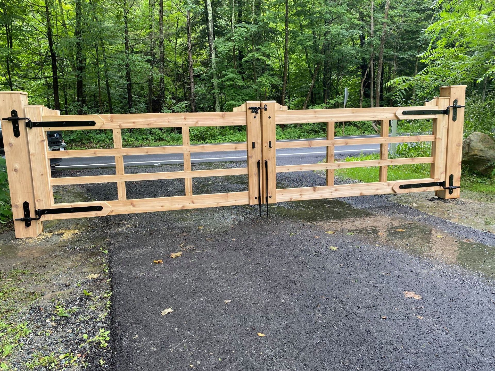 Wooden gate with black hinges, closed, over a paved driveway, forest background.