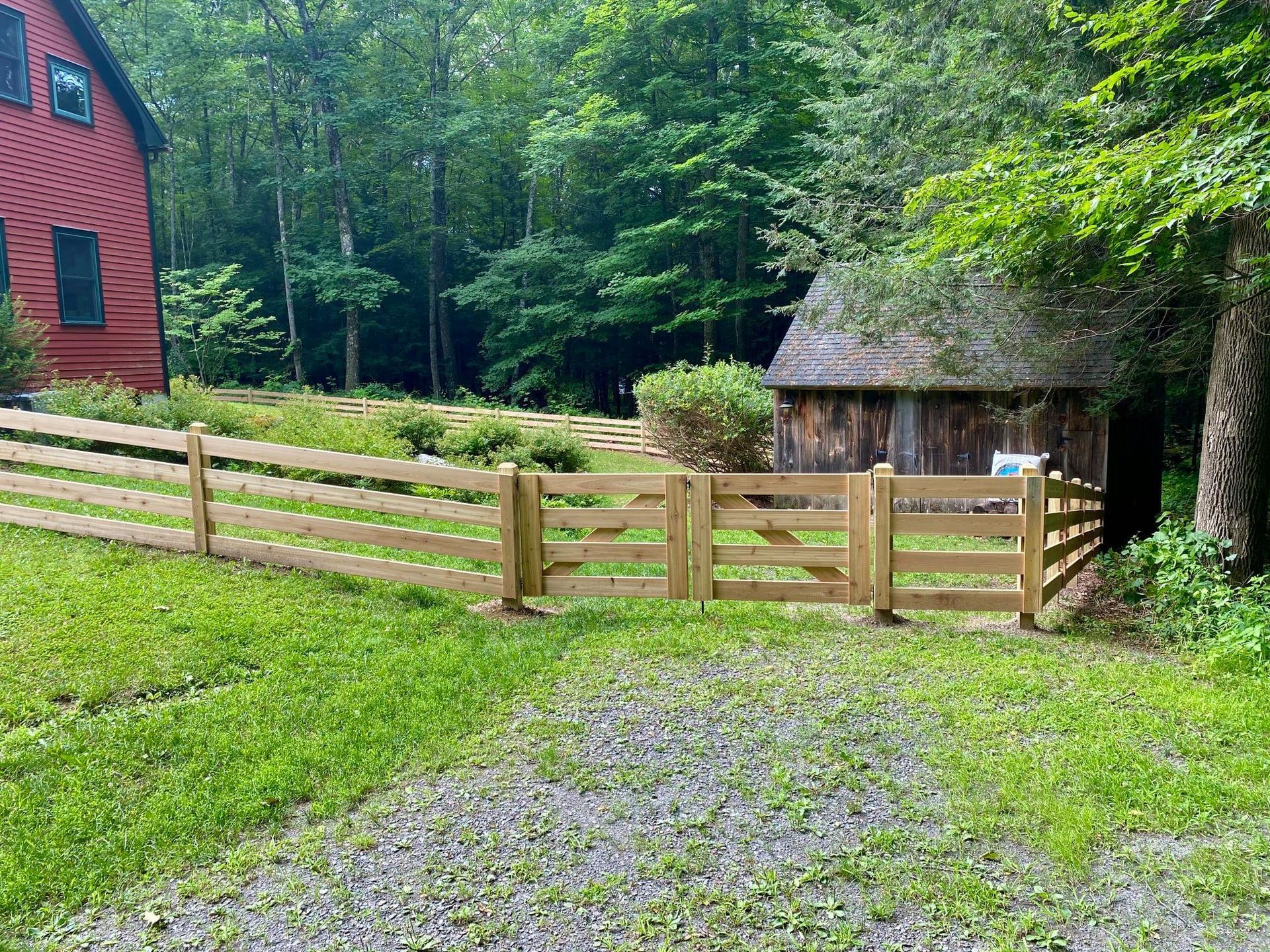 Wooden fence and gates in front of a red house and shed, with green grass and trees in the background.