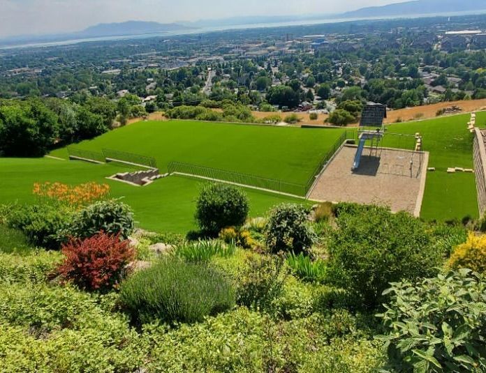 Green hillside overlooking a sunlit valley with lawns, trees, and a few buildings in the distance