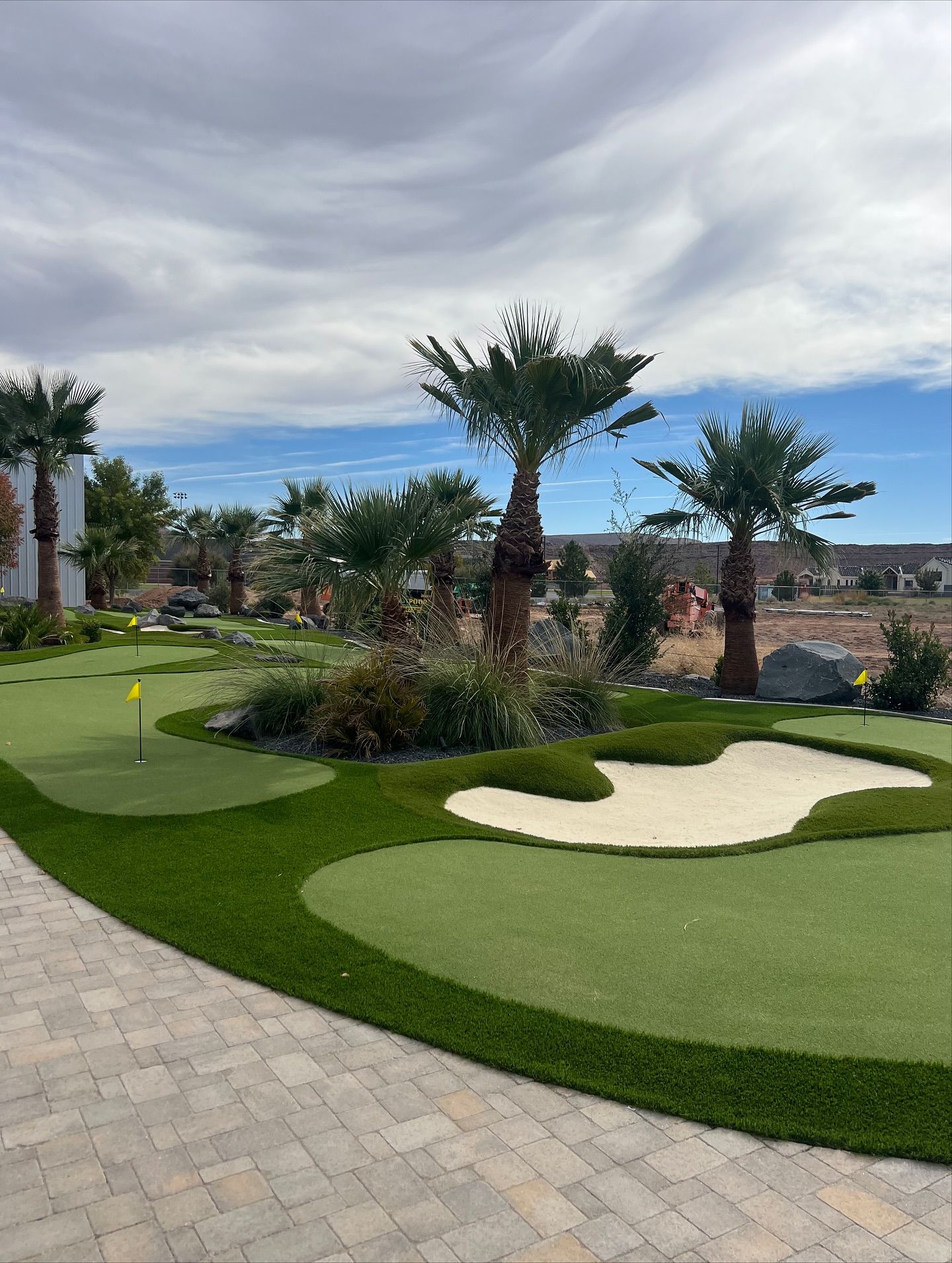 Landscaped mini golf course with palm trees, green turf, sand traps, and cloudy sky
