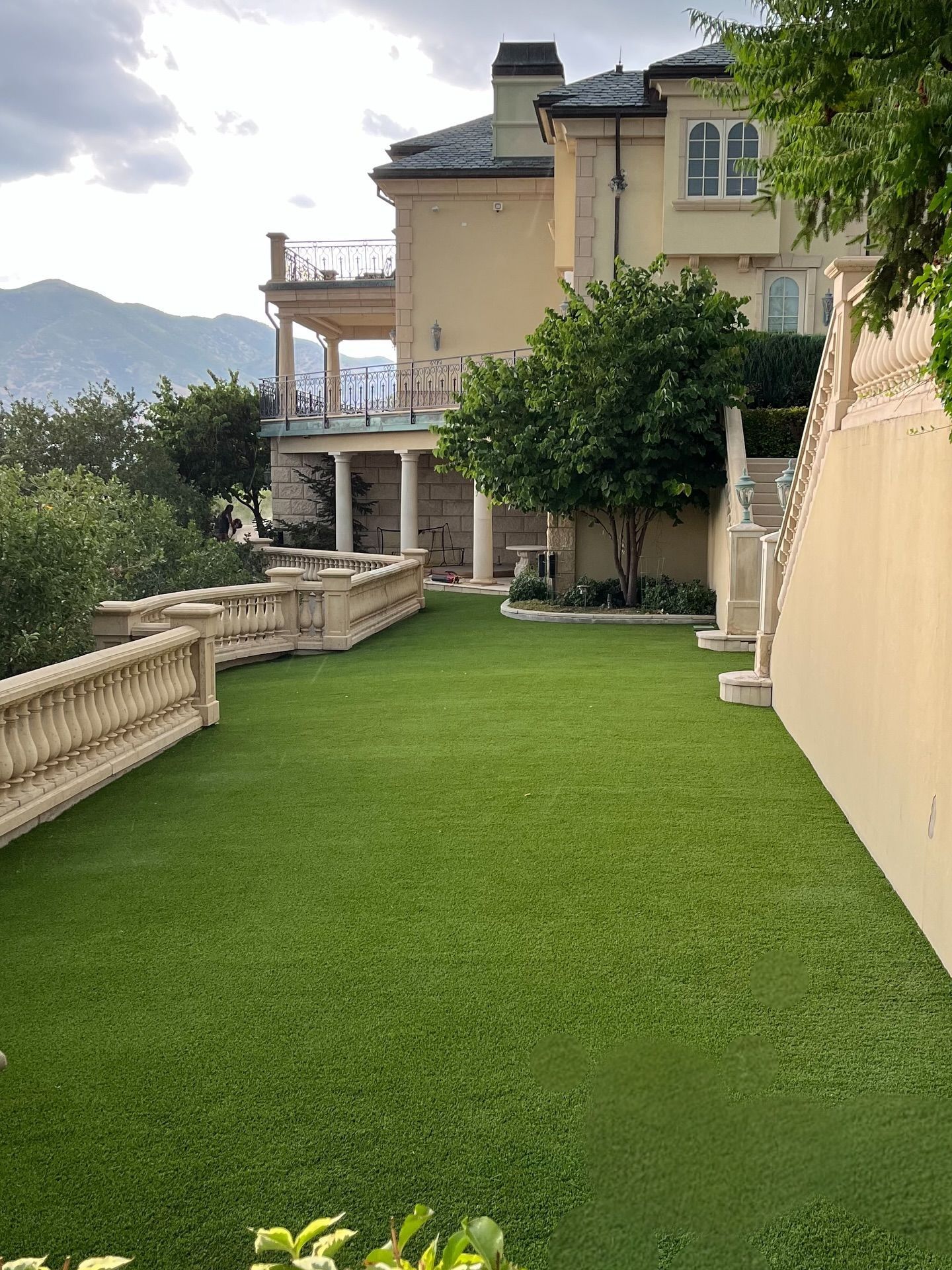 Lush green lawn beside a beige villa with balconies, trees, and a mountain view at sunset.