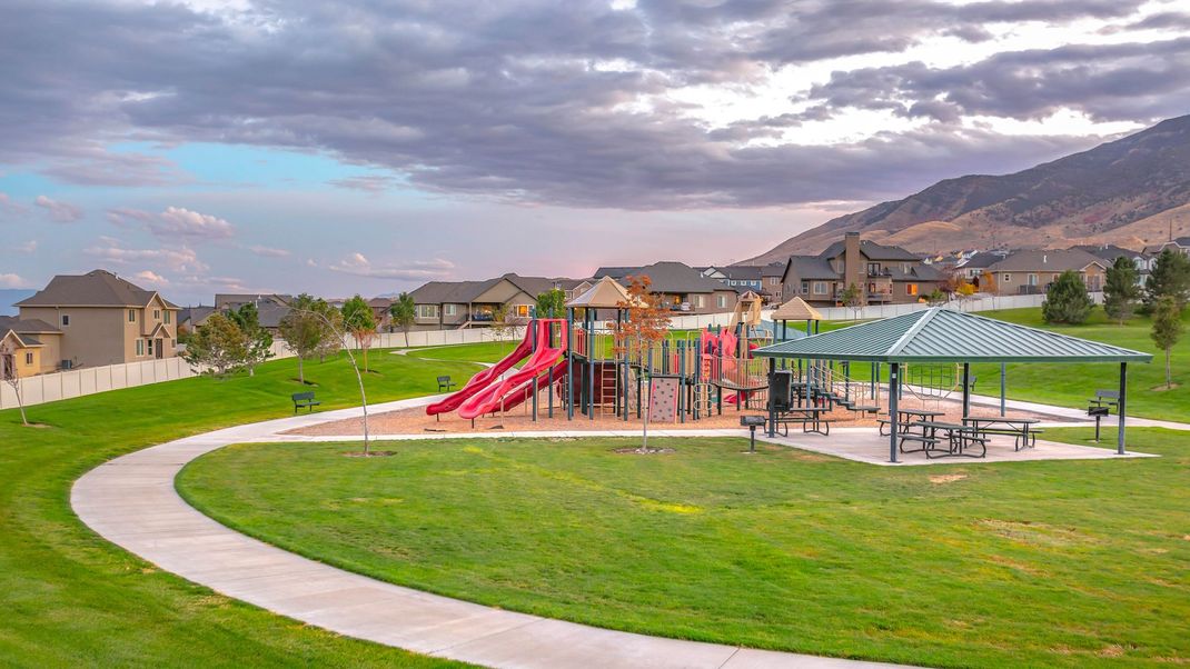 Playground with red slides beside a curving path, green lawn, and mountains under a cloudy sky