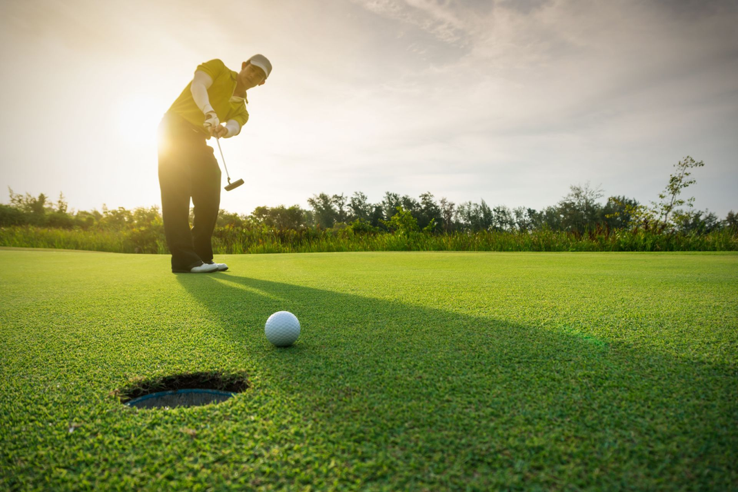 Golfer putting on a sunlit green near the hole, with the ball just short of the cup.