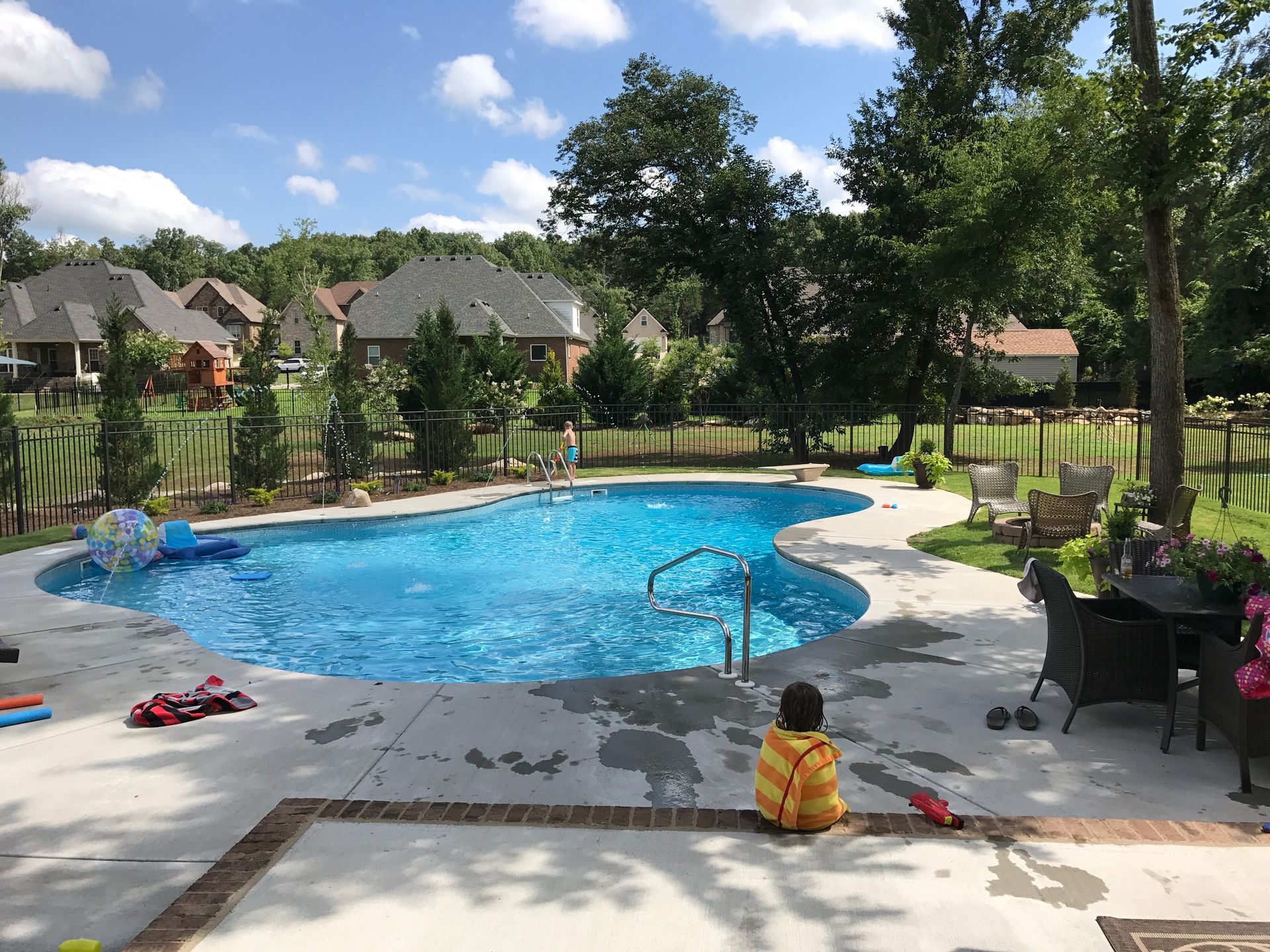 A child in a yellow hooded towel sits poolside on a sunny day in a backyard with a blue swimming pool and patio furniture.