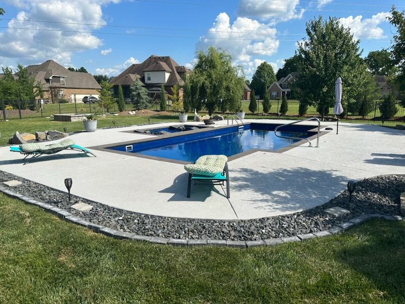 A backyard swimming pool with a spa, lounge chairs, and a patio surrounded by a decorative rock border and lawn.
