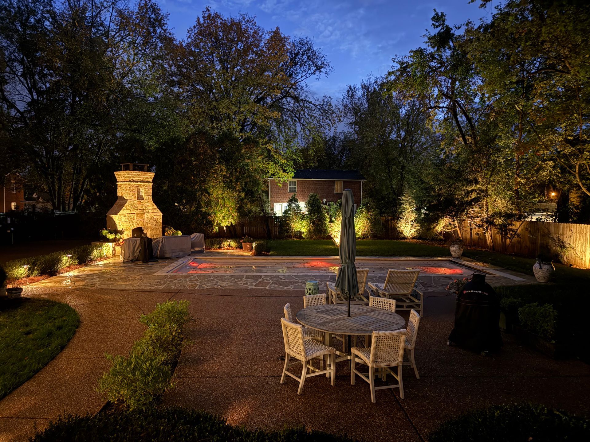 An illuminated backyard patio at twilight with a stone fireplace, dining table, and swimming pool in the background.