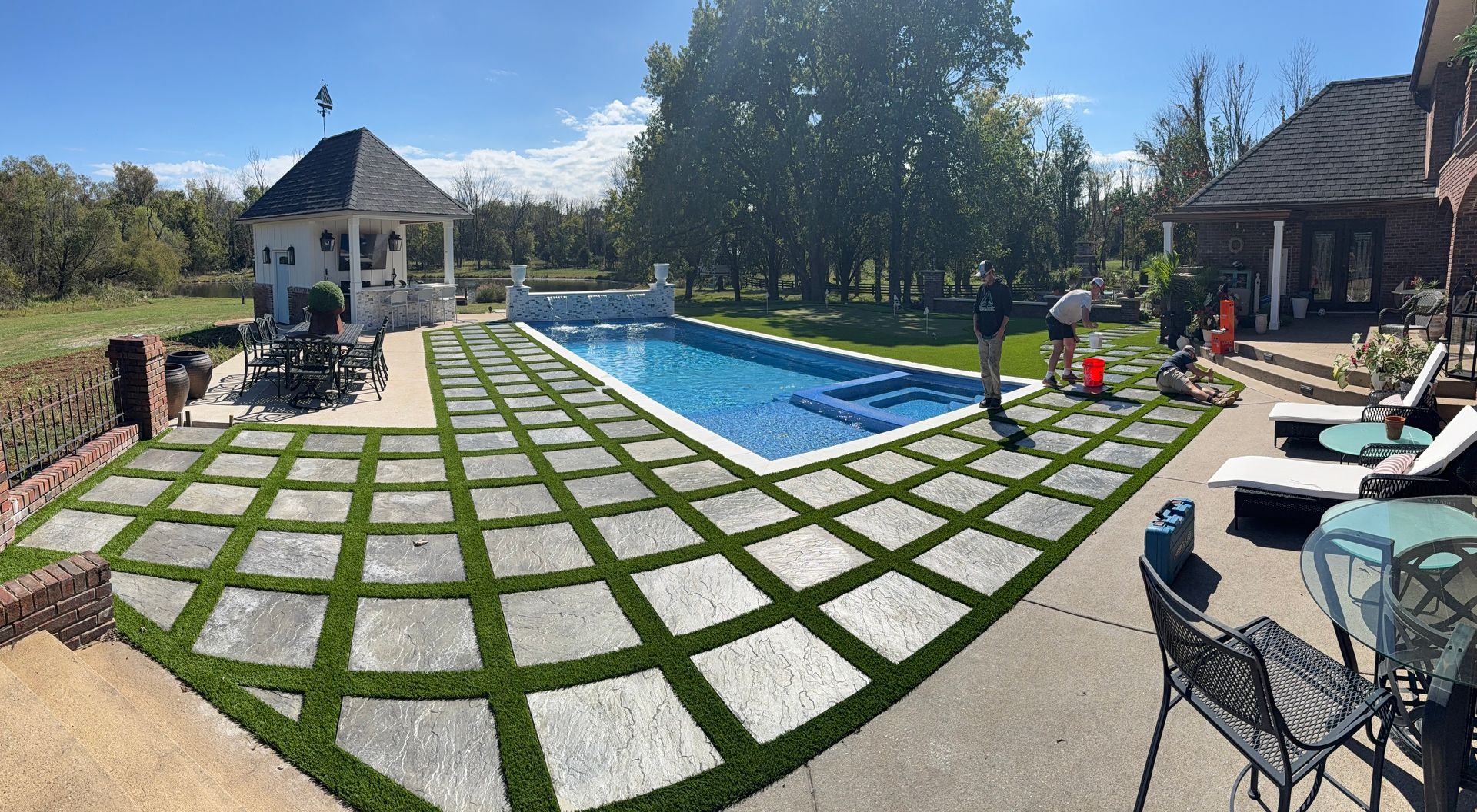 A backyard pool area featuring a rectangular pool surrounded by square stone pavers with grass inlays and a gazebo.