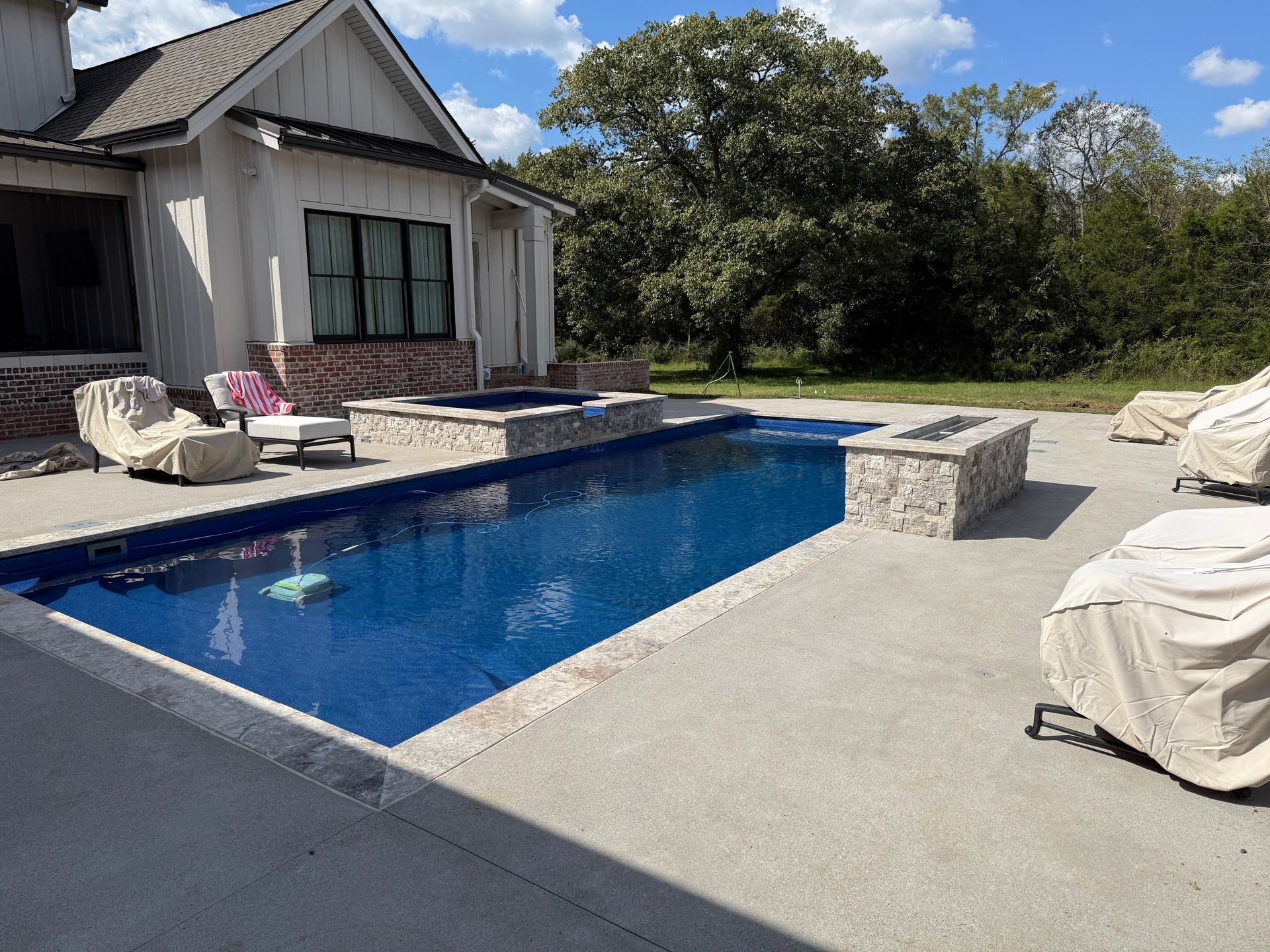 A backyard with a rectangular blue swimming pool, stone hot tub, fire feature, and outdoor lounge furniture on a patio.