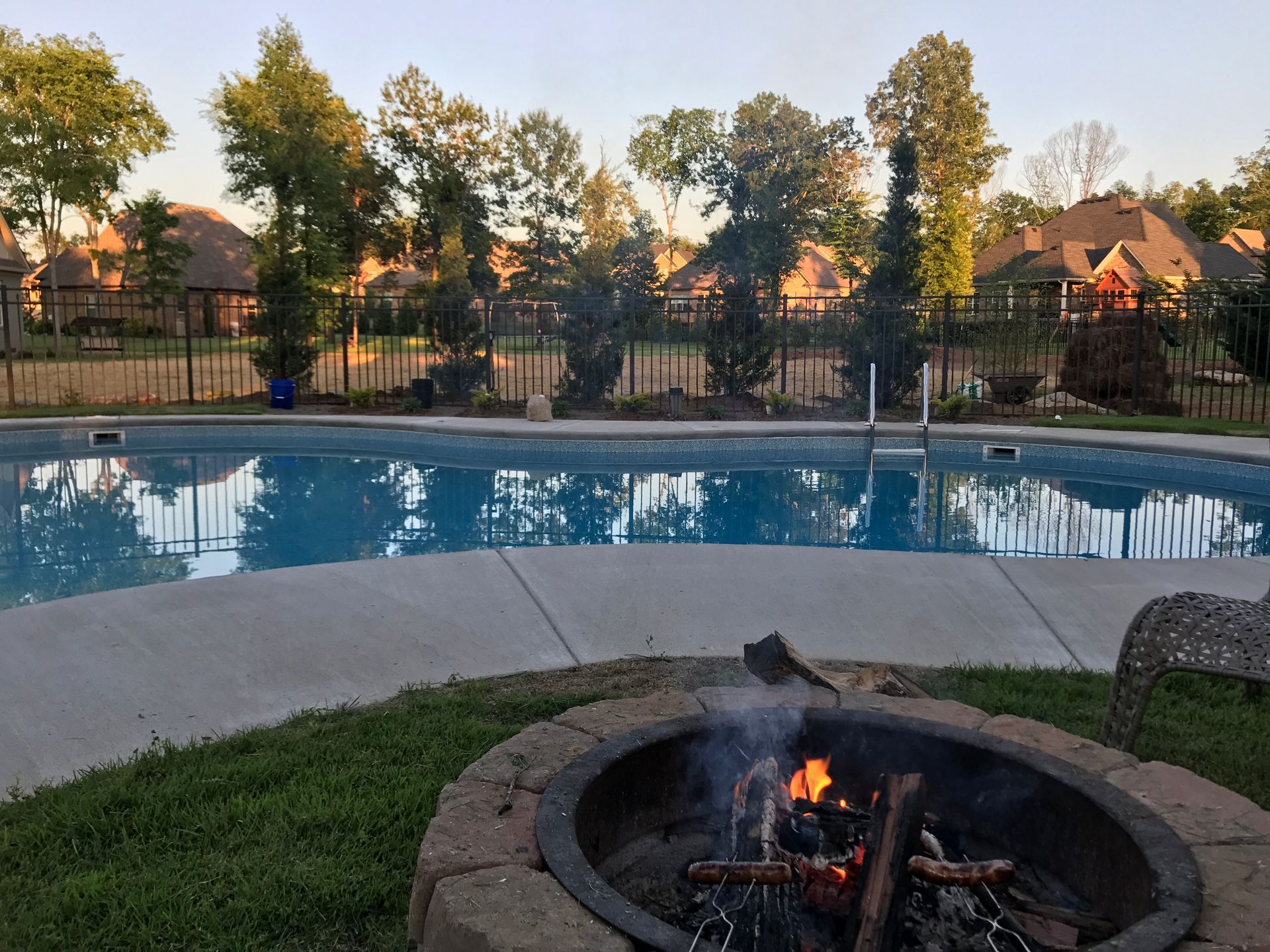A backyard fire pit with a glowing fire next to a blue swimming pool at dusk, with houses and trees in the background.