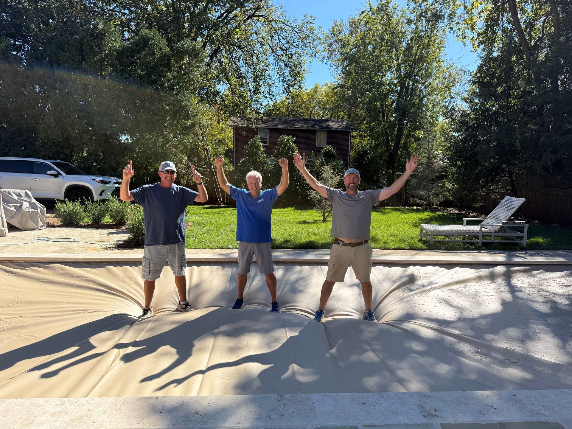 Three people stand with arms raised on a large, tan, taut pool safety cover in a sunny, green backyard.