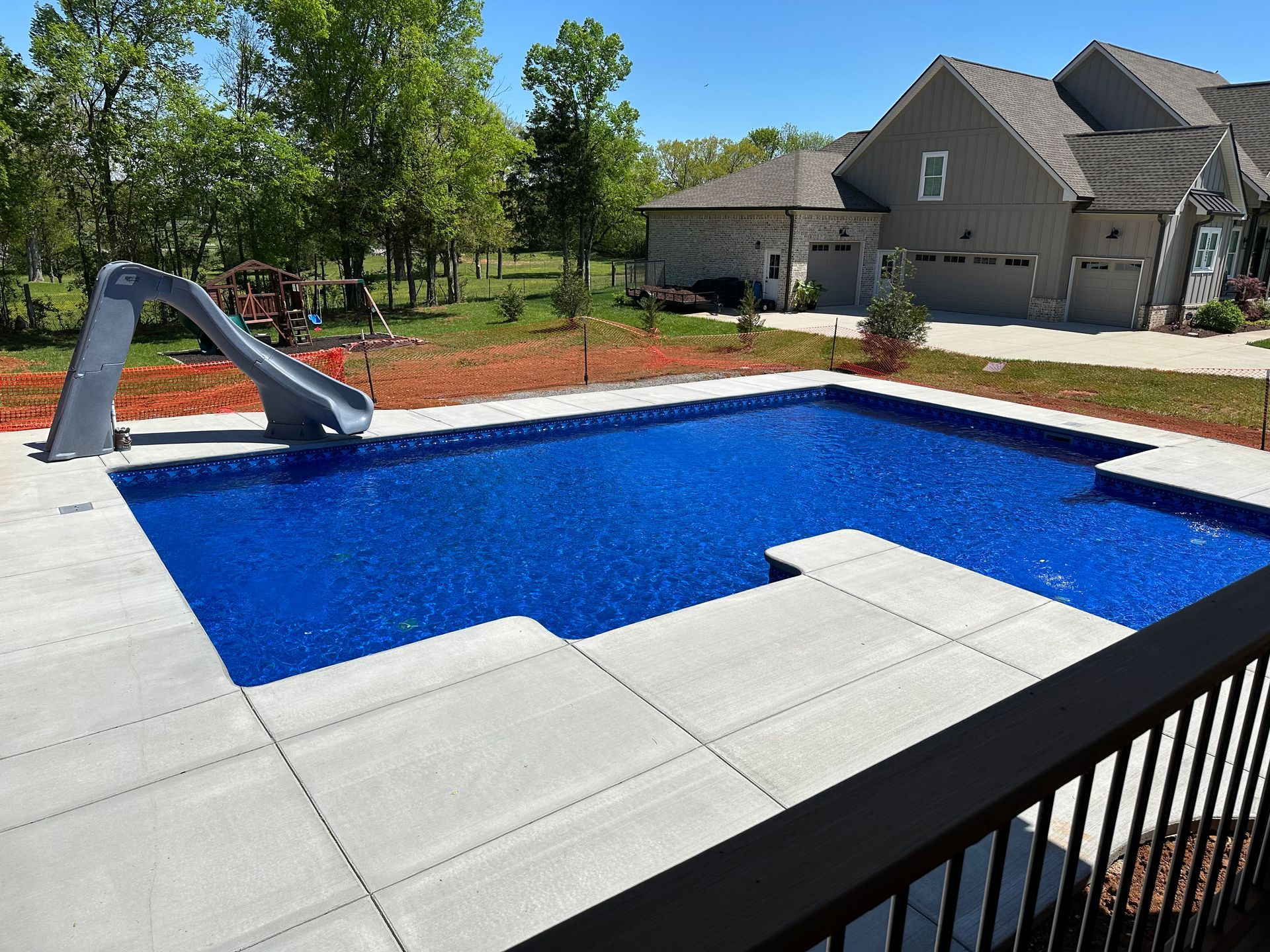 A bright blue rectangular swimming pool with a grey slide and concrete patio, set in a backyard near a large house.