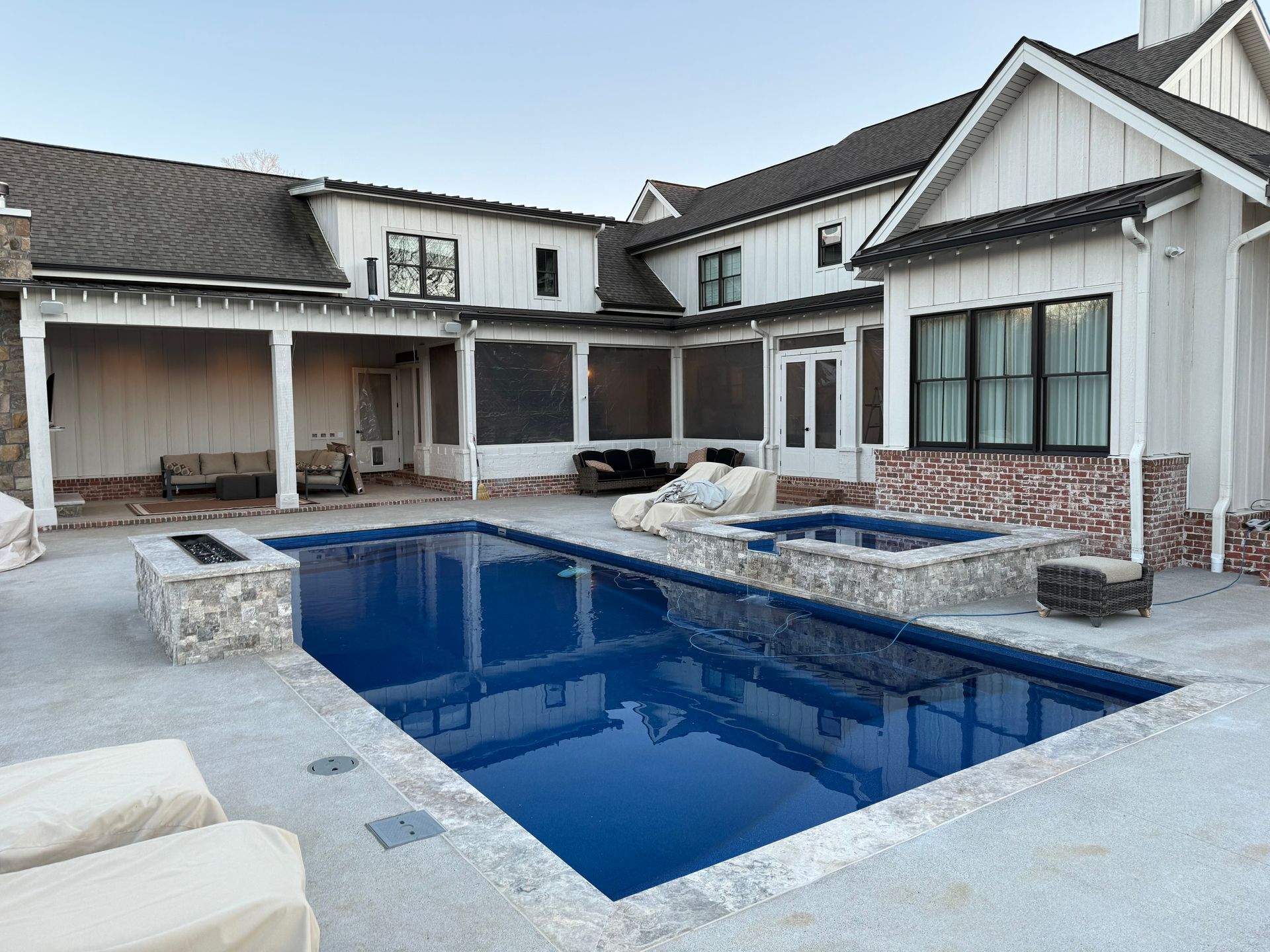 A modern white farmhouse with a patio, swimming pool, and stone fire pit in a backyard setting at sunset.