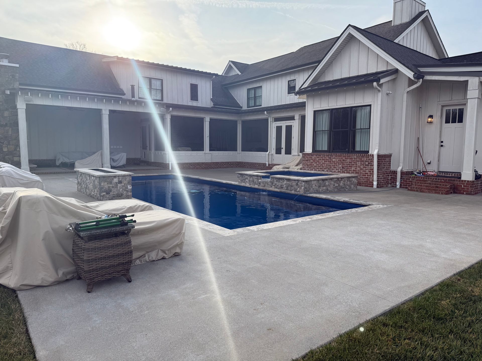A backyard with a rectangular pool and stone fire pit near a white farmhouse-style home with a covered patio.