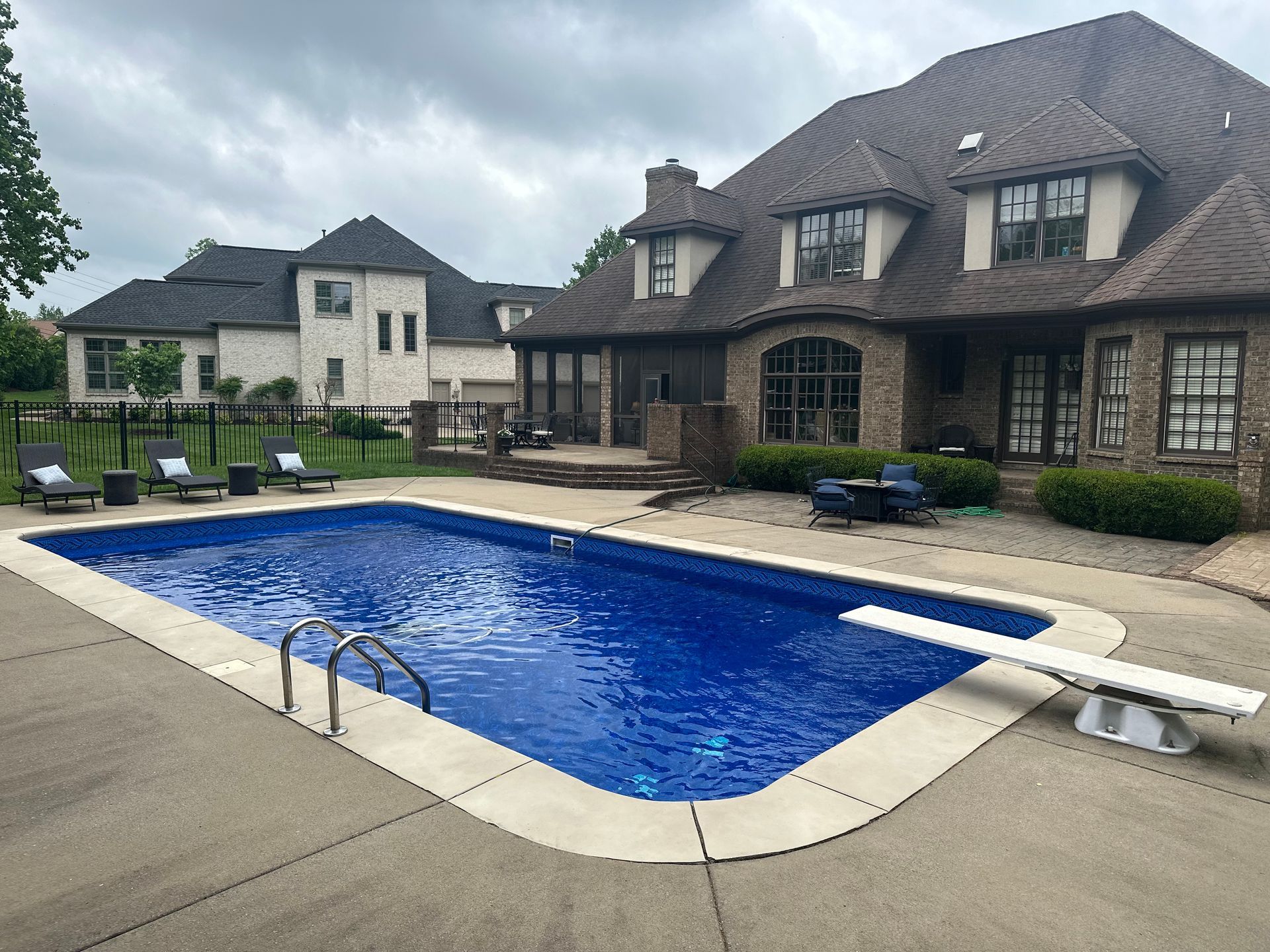 A large backyard features a rectangular blue swimming pool with a diving board next to a two-story brick and stone house.
