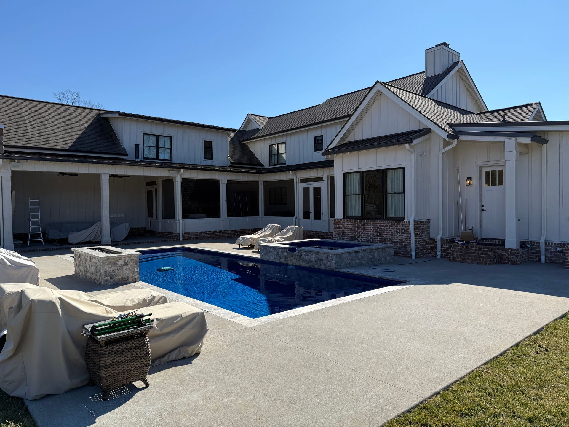 A white modern farmhouse with a pool, hot tub, patio furniture, and a stone fire pit on a sunny day.