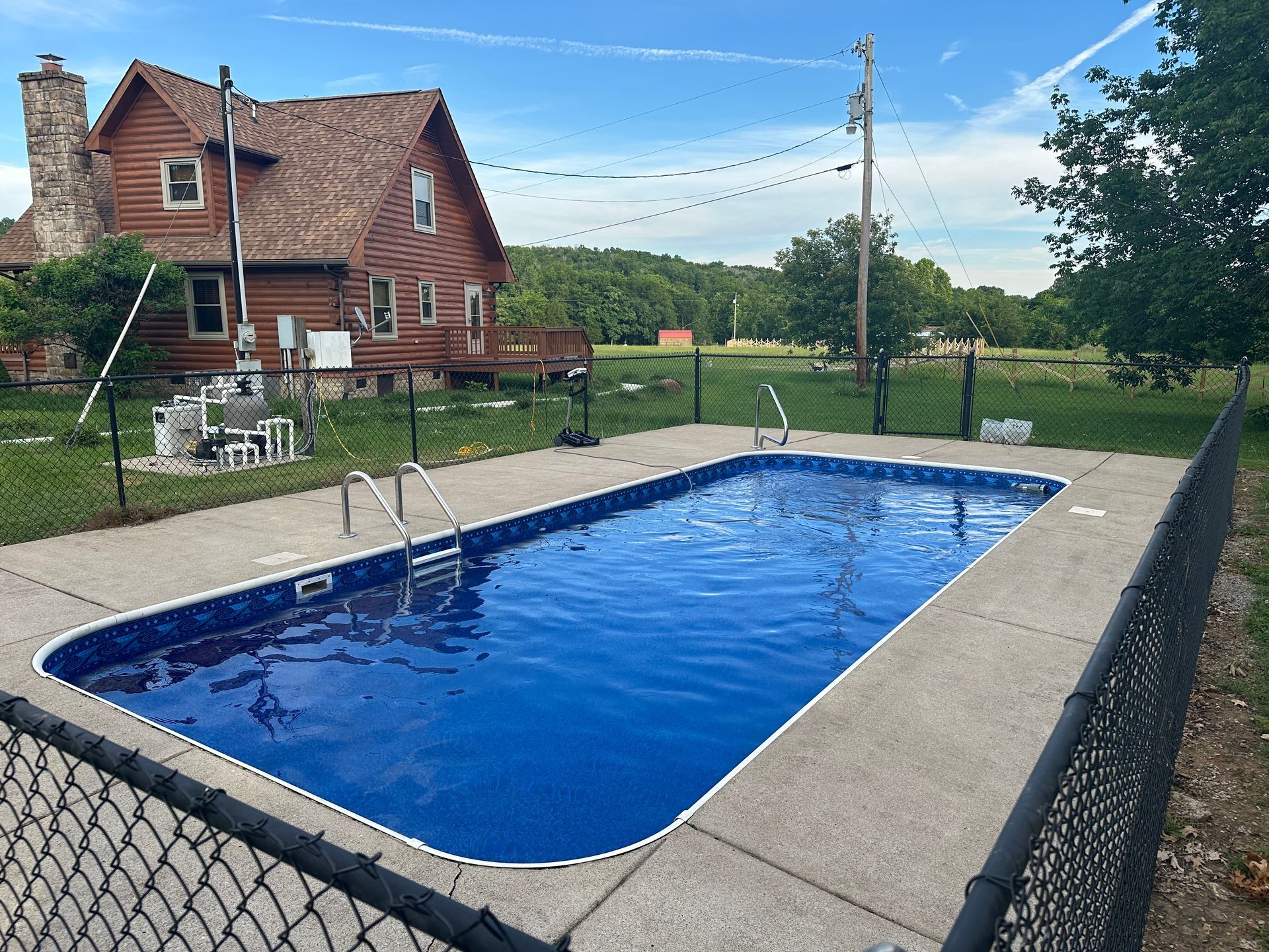 A rectangular swimming pool with bright blue water sits in a backyard next to a rustic log house under a sunny sky.