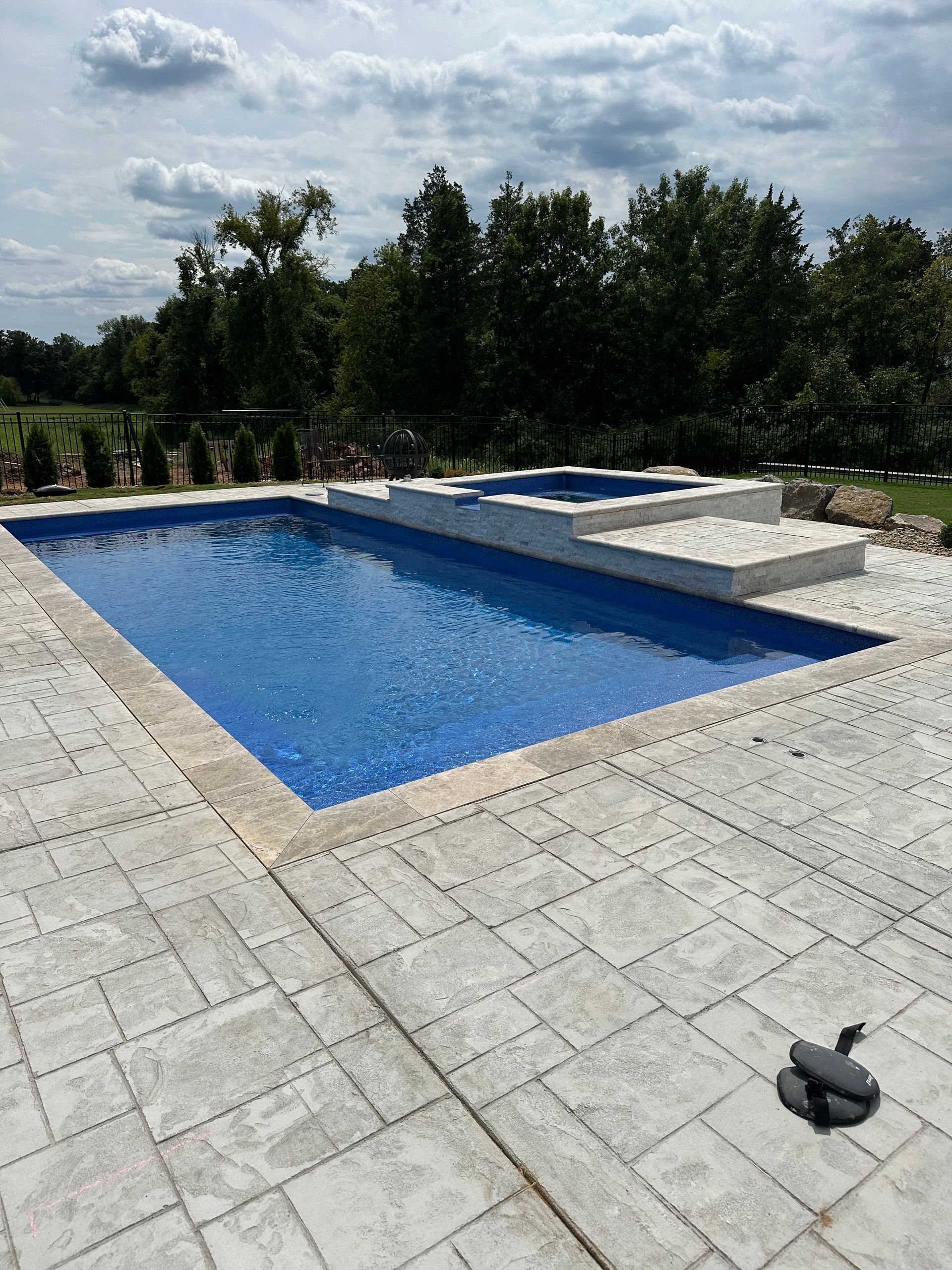 A rectangular blue swimming pool with an attached raised spa, surrounded by light gray stone pavers under a cloudy sky.