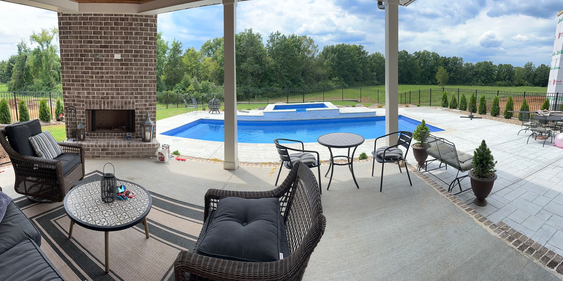 A covered patio with brick fireplace furniture facing an outdoor swimming pool and lush green landscape.