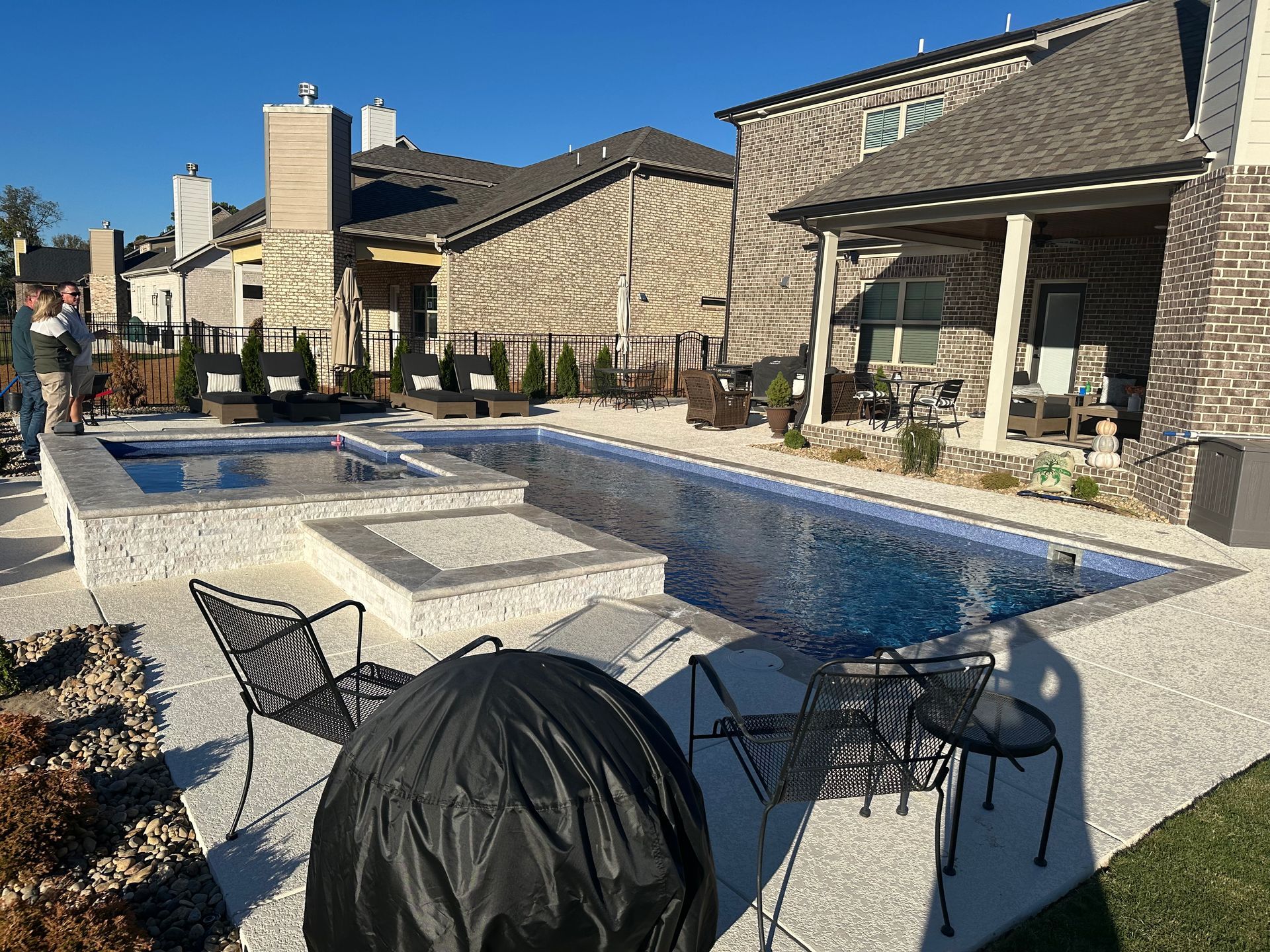 A backyard swimming pool and hot tub next to a stone house with patio furniture on a sunny day.