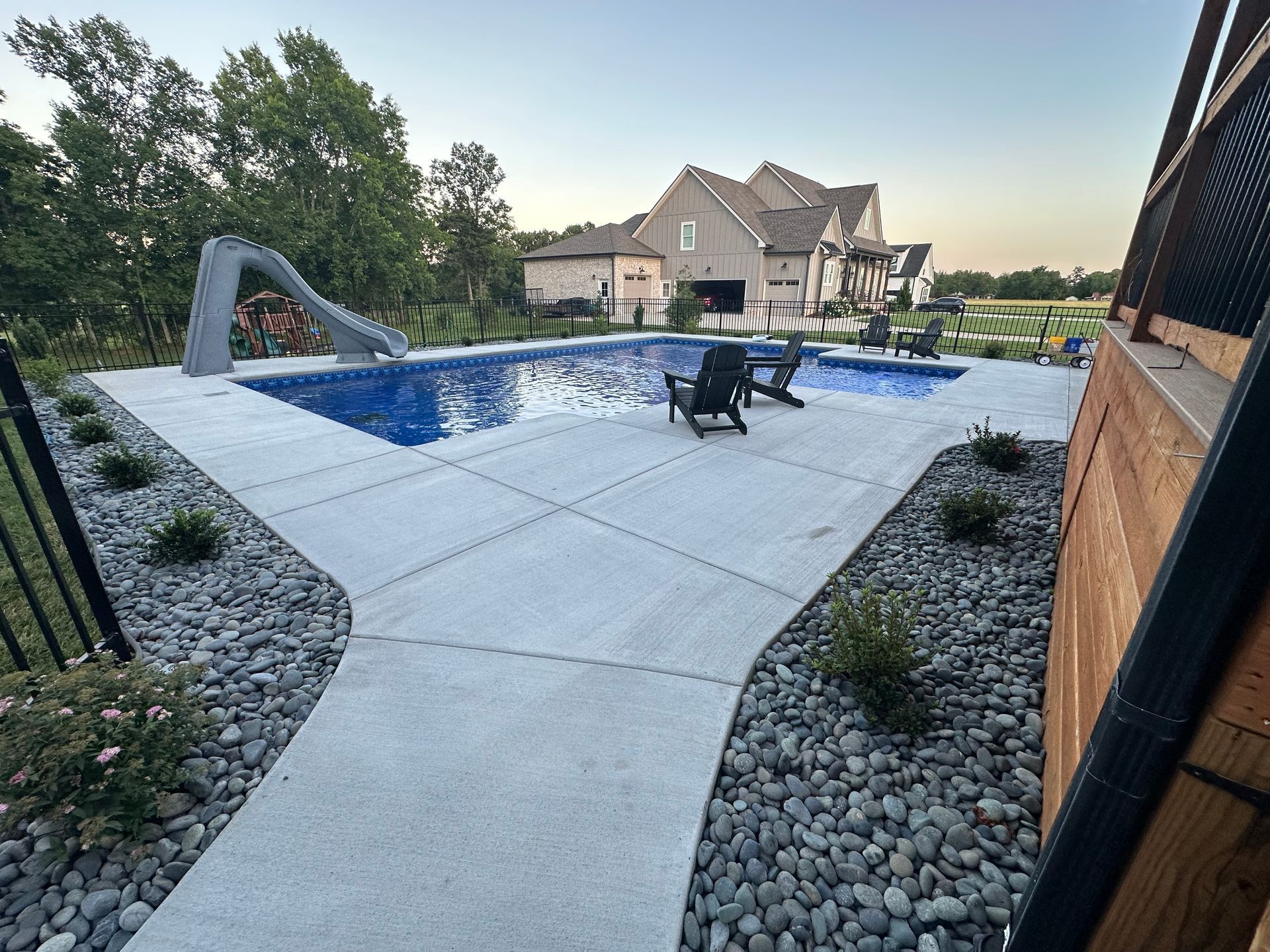 A swimming pool with a slide, surrounded by a concrete patio and landscaping rock, with a house in the background.
