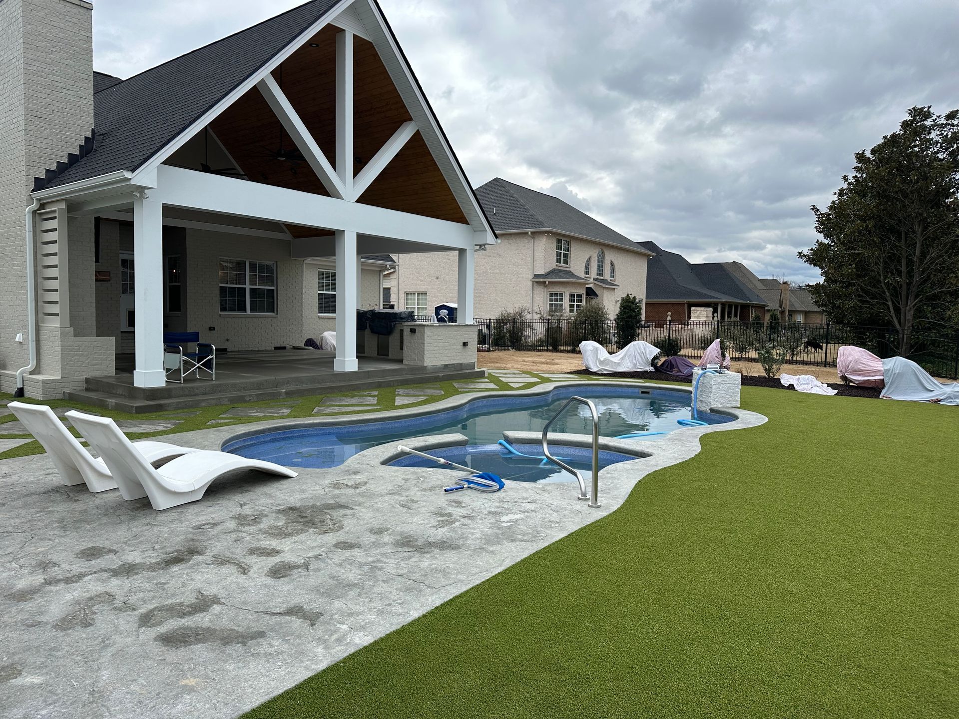 A backyard features a swimming pool with lounge chairs on a stone deck, adjacent to a house with a covered patio.