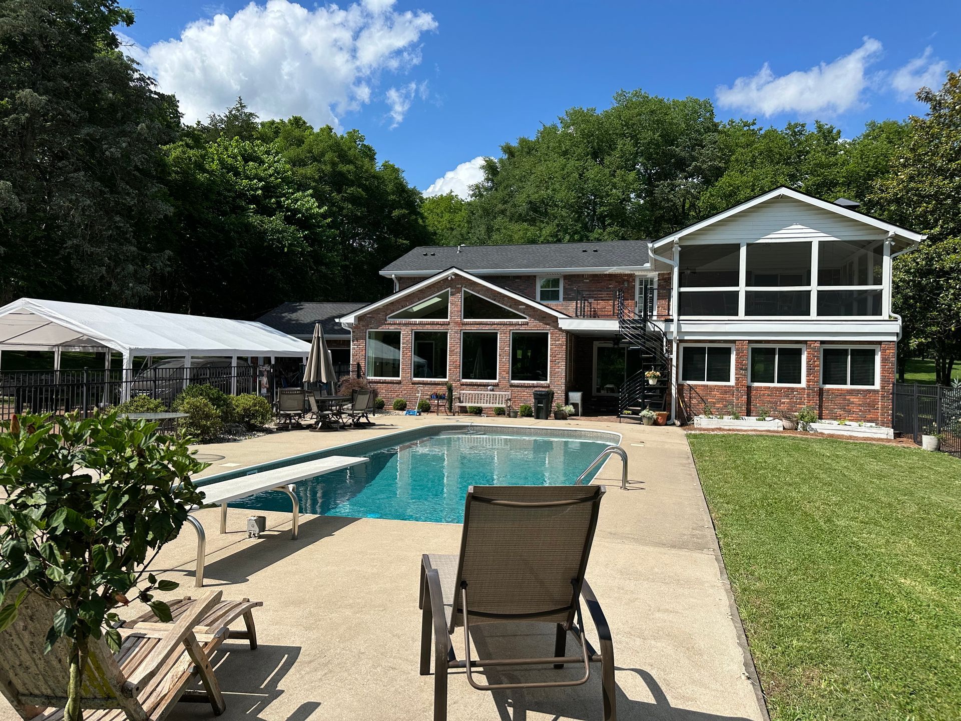 A sunny backyard with a rectangular swimming pool, lounge chairs, a white tent, and a two-story brick and siding house.