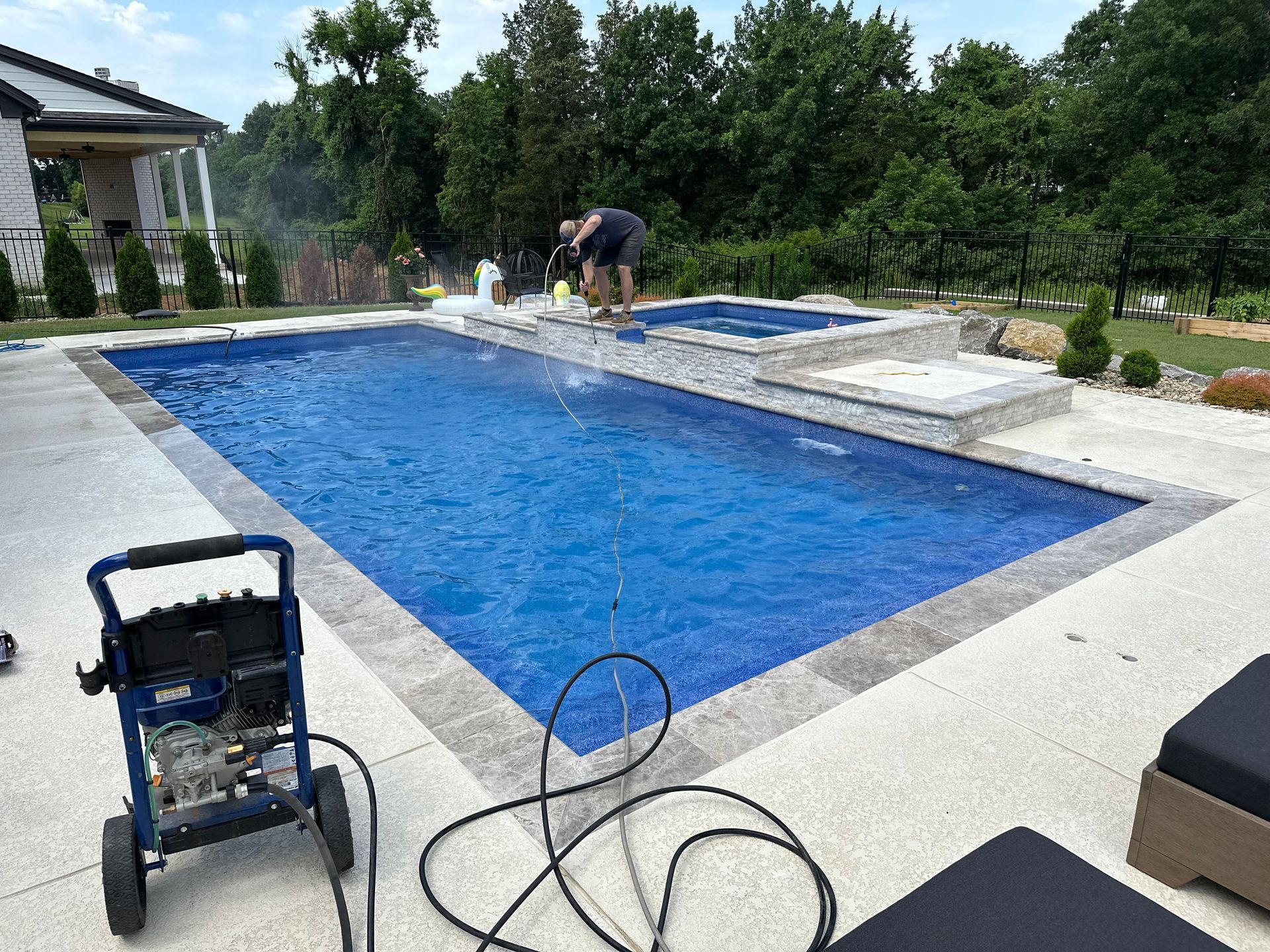 A person pressure washes the stone coping of a rectangular backyard pool with a blue-tiled interior.