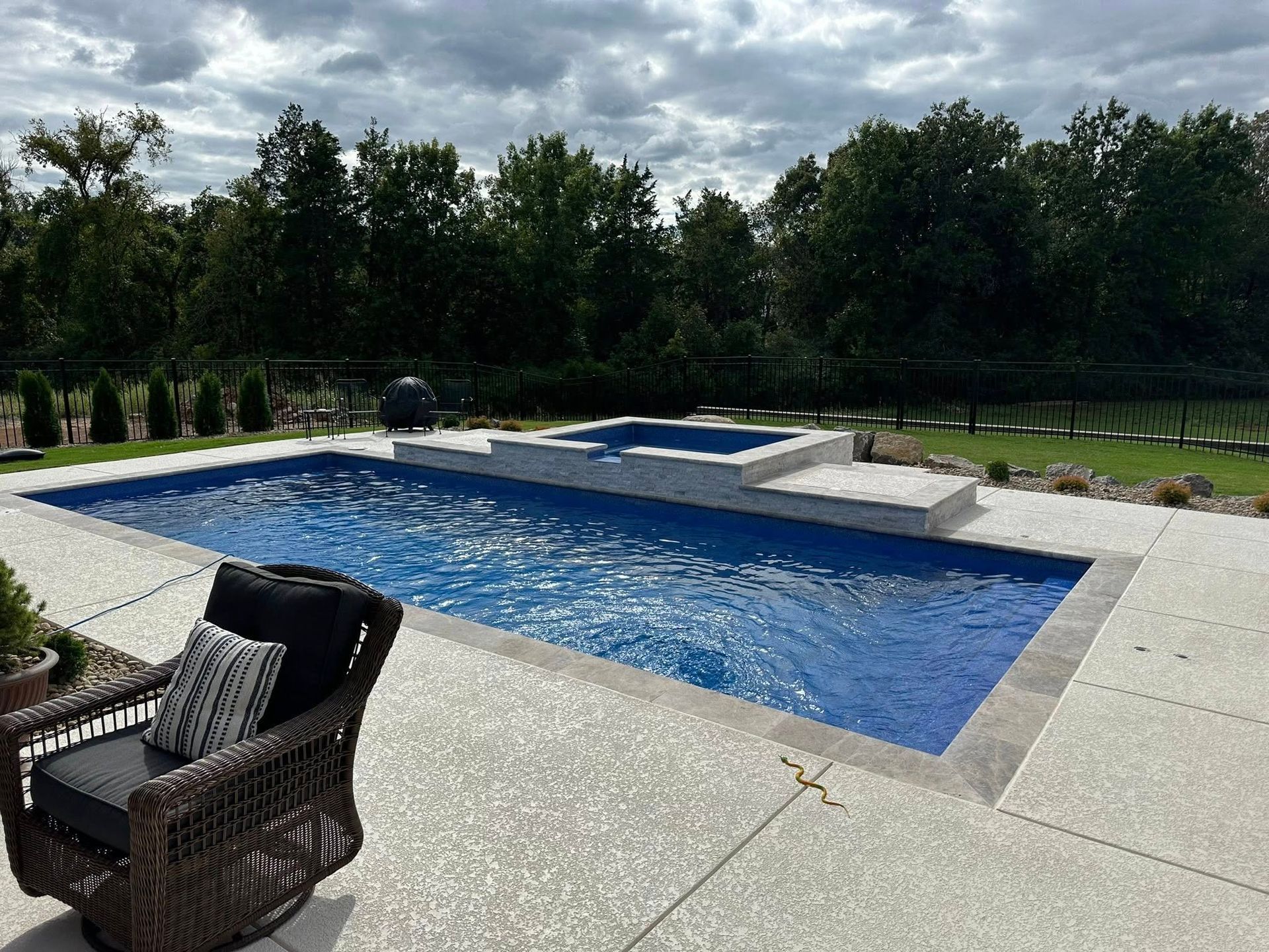 Swimming pool with attached spa, surrounded by a patio, trees and a lounge chair.