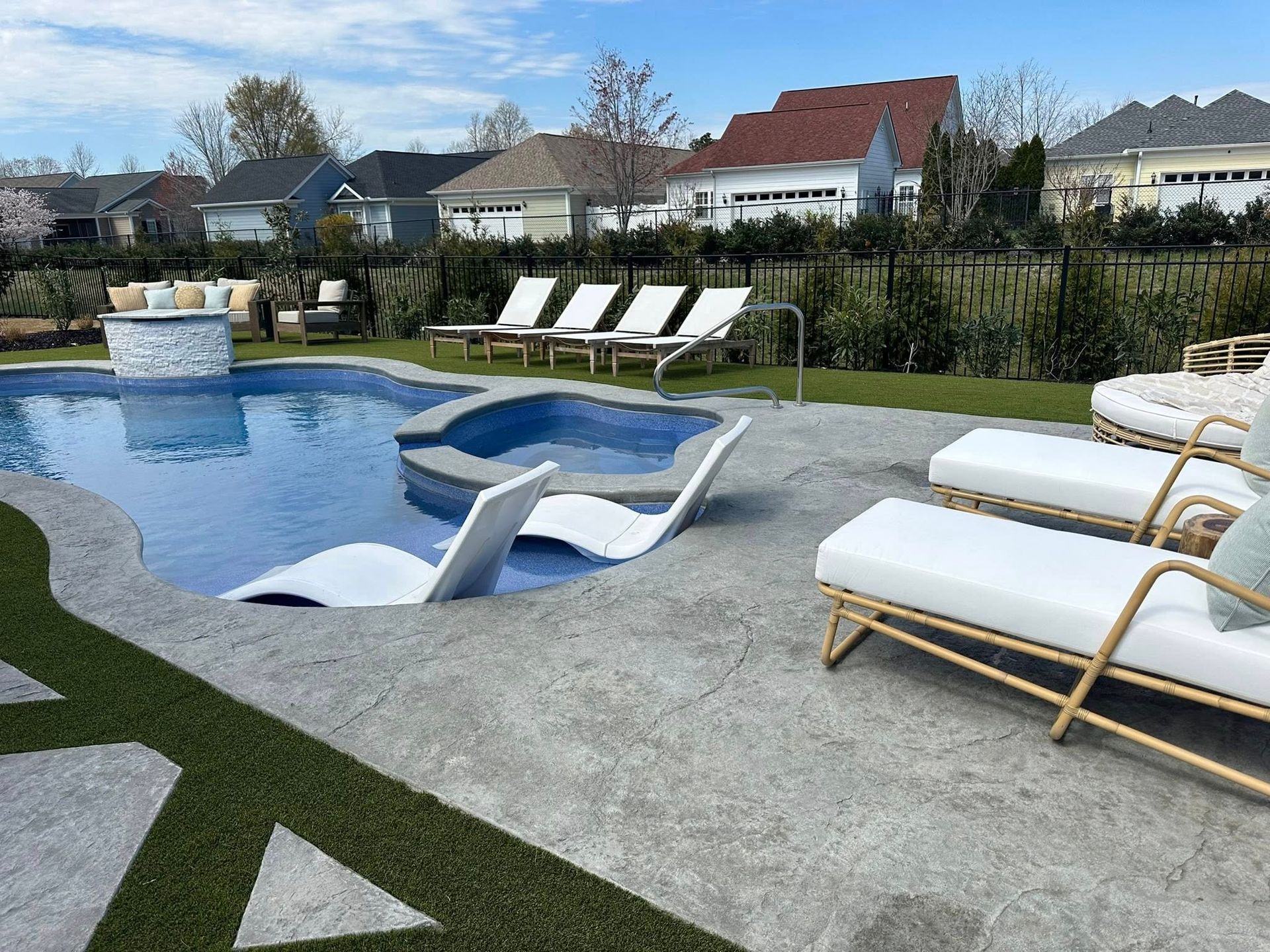 Outdoor pool area with lounge chairs, hot tub, and fire pit. Houses in the background under a blue sky.